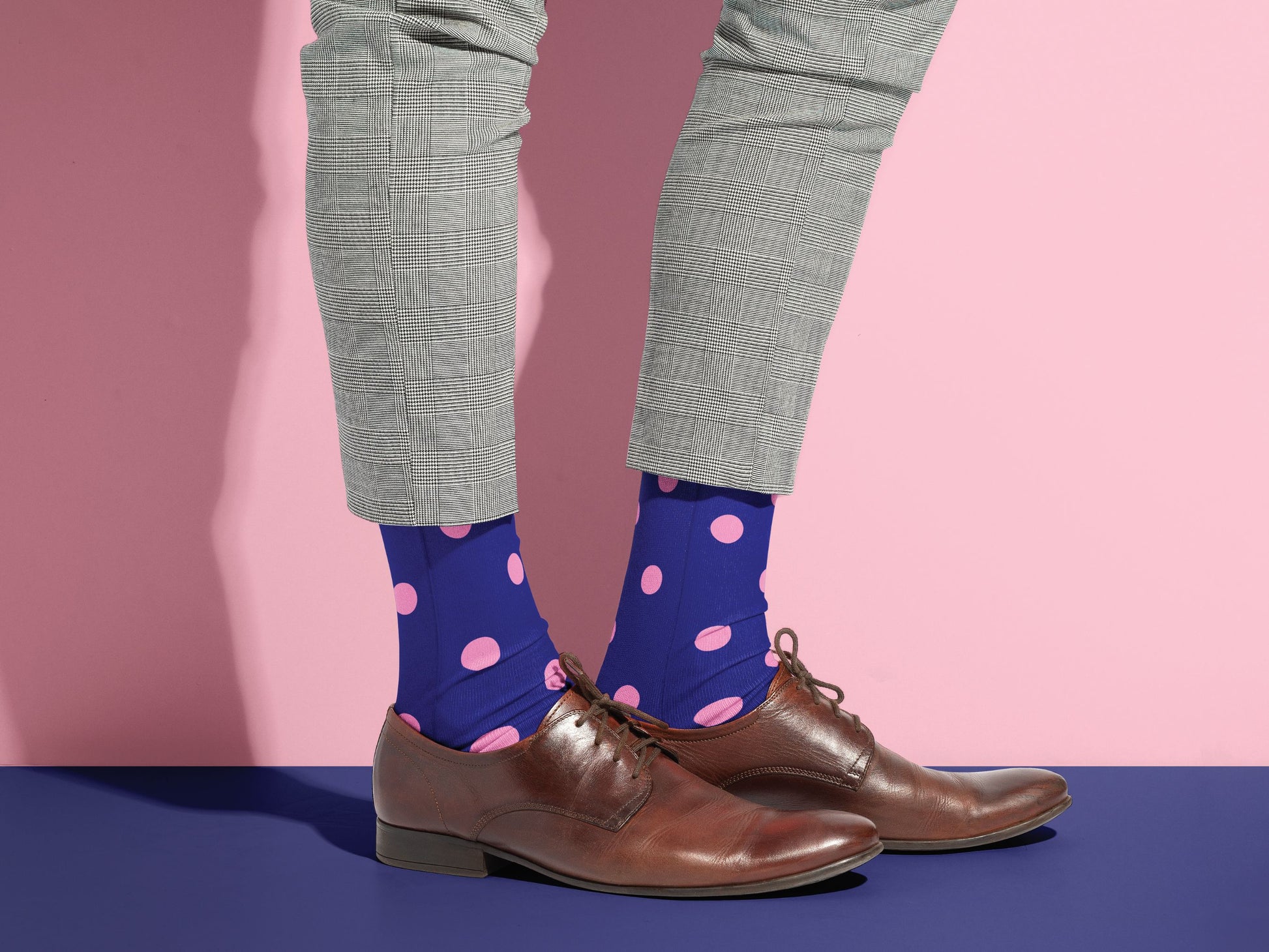 Person wearing blue socks with pink polka dots, gray pants, and brown shoes on a pink and blue gradient background.