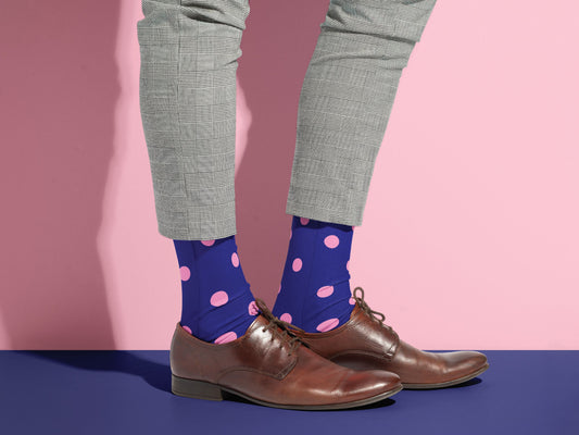 Person wearing blue socks with pink polka dots, gray pants, and brown shoes on a pink and blue gradient background.
